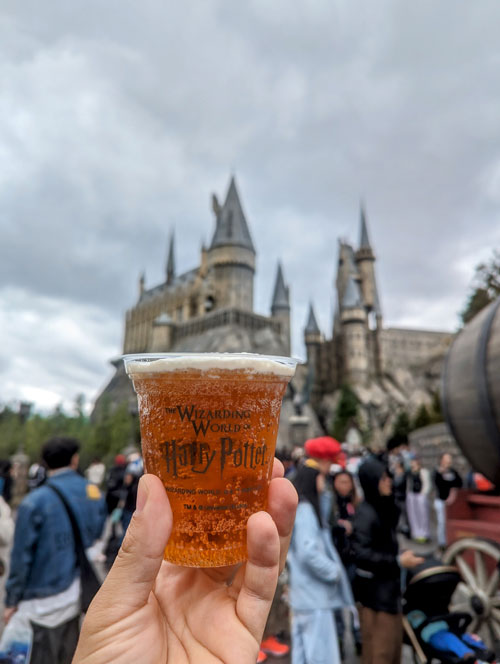 Mano sosteniendo una cerveza de mantequilla en un vaso de recuerdo frente al castillo de Hogwarts.