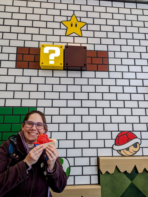 La esposa de Pablo, autor de Vamos Pa' Japón, posando con un recuerdo de Mario frente a una pared decorada con ladrillos, un bloque de interrogación y un caparazón rojo.