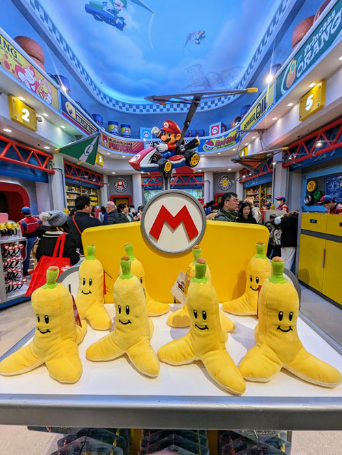 Peluches de plátano de Mario Kart en una tienda de Super Nintendo World, con la figura de Mario en su kart al fondo.