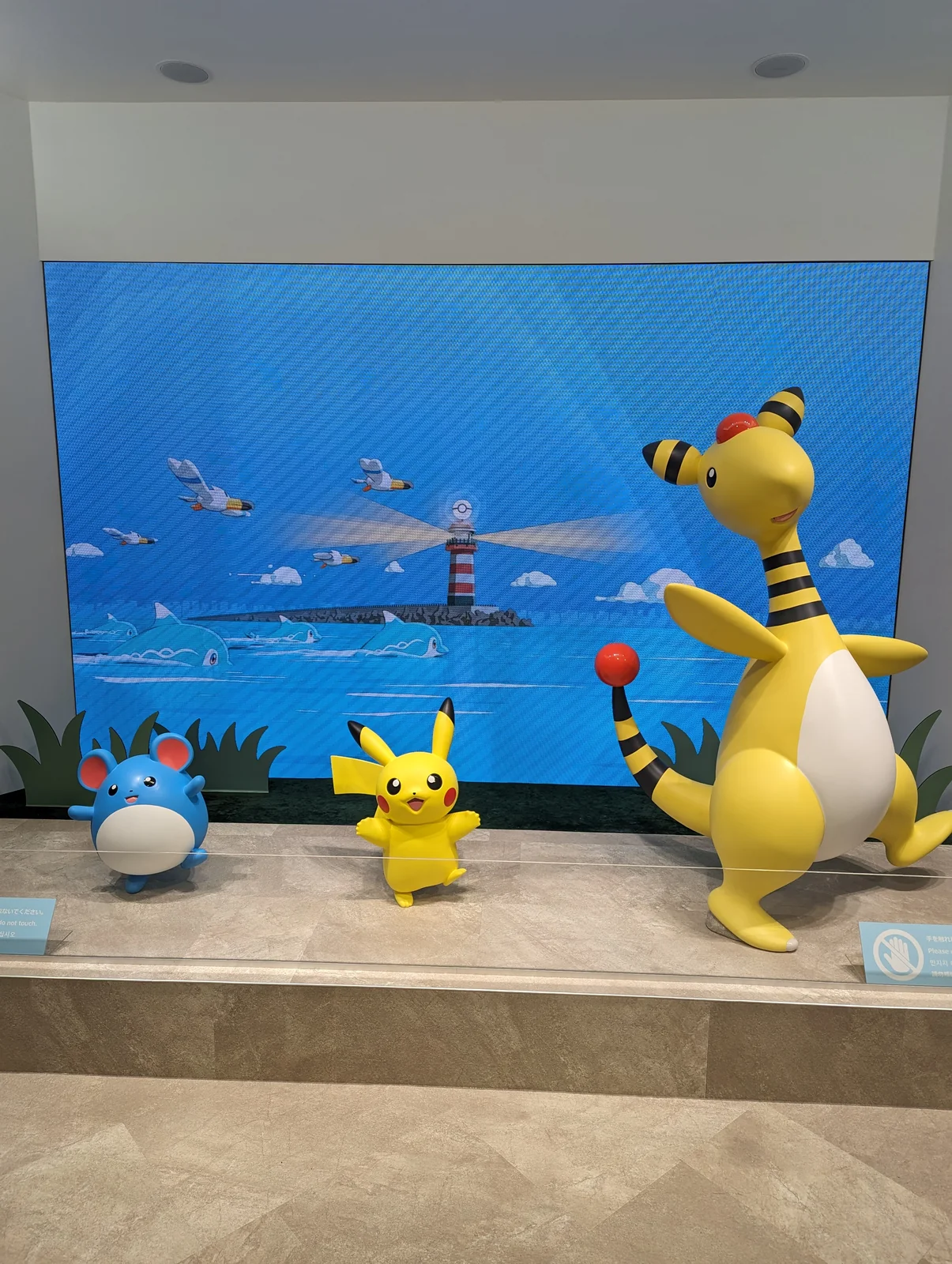 Ampharos, Pikachu y Marill frente a un mural de la bahía con un faro en el Pokémon Center Tokyo-Bay